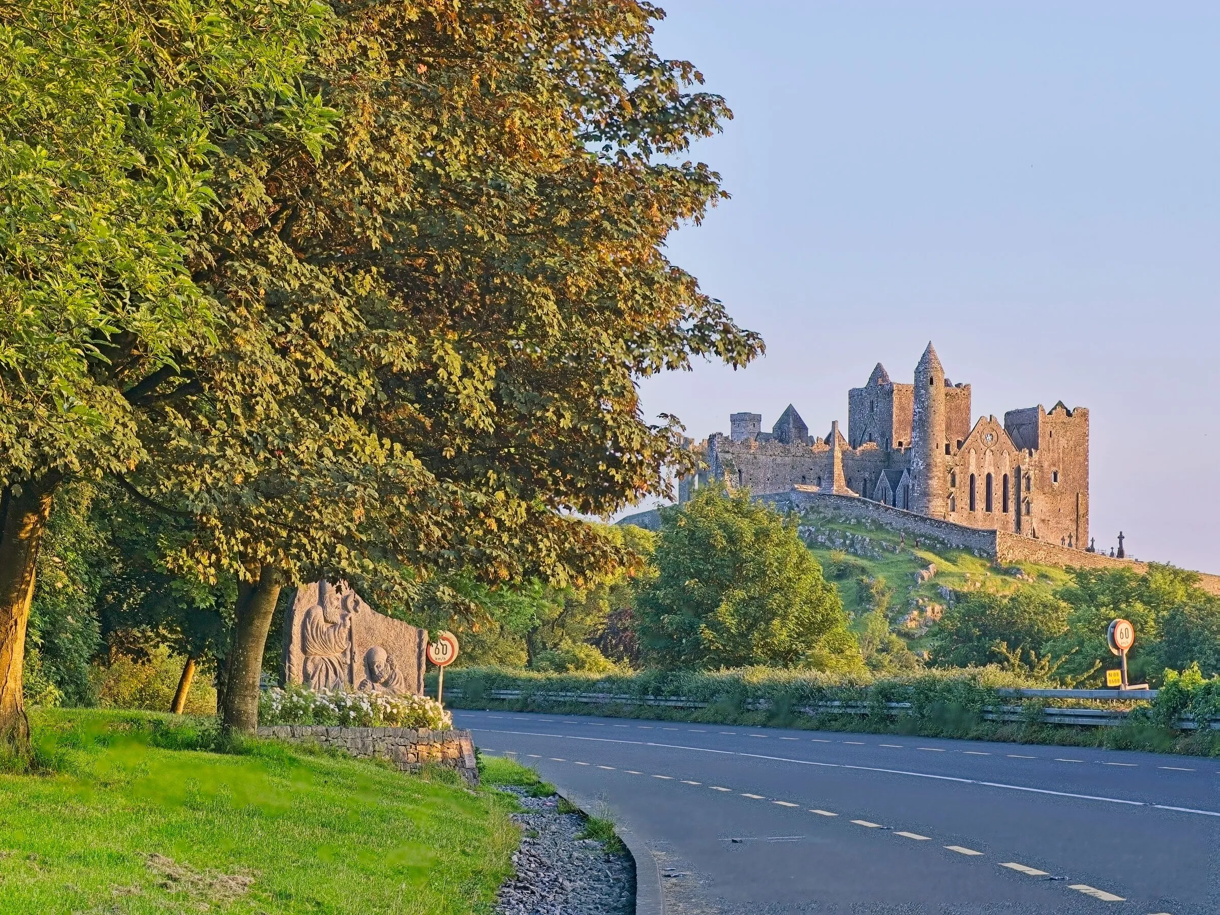 The Rock of Cashel on its ridge above the road into Cashel, lit gold by low sun, with trees in the foreground