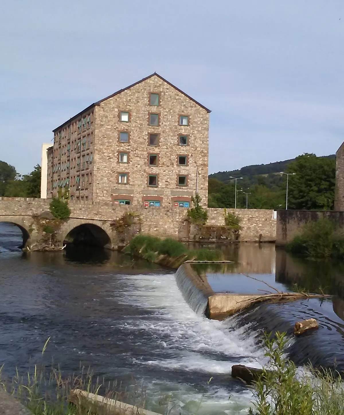 The River Suir at Clonmel running white over the weir beside the arches of the Old Bridge, with a stone mill building on the bank behind