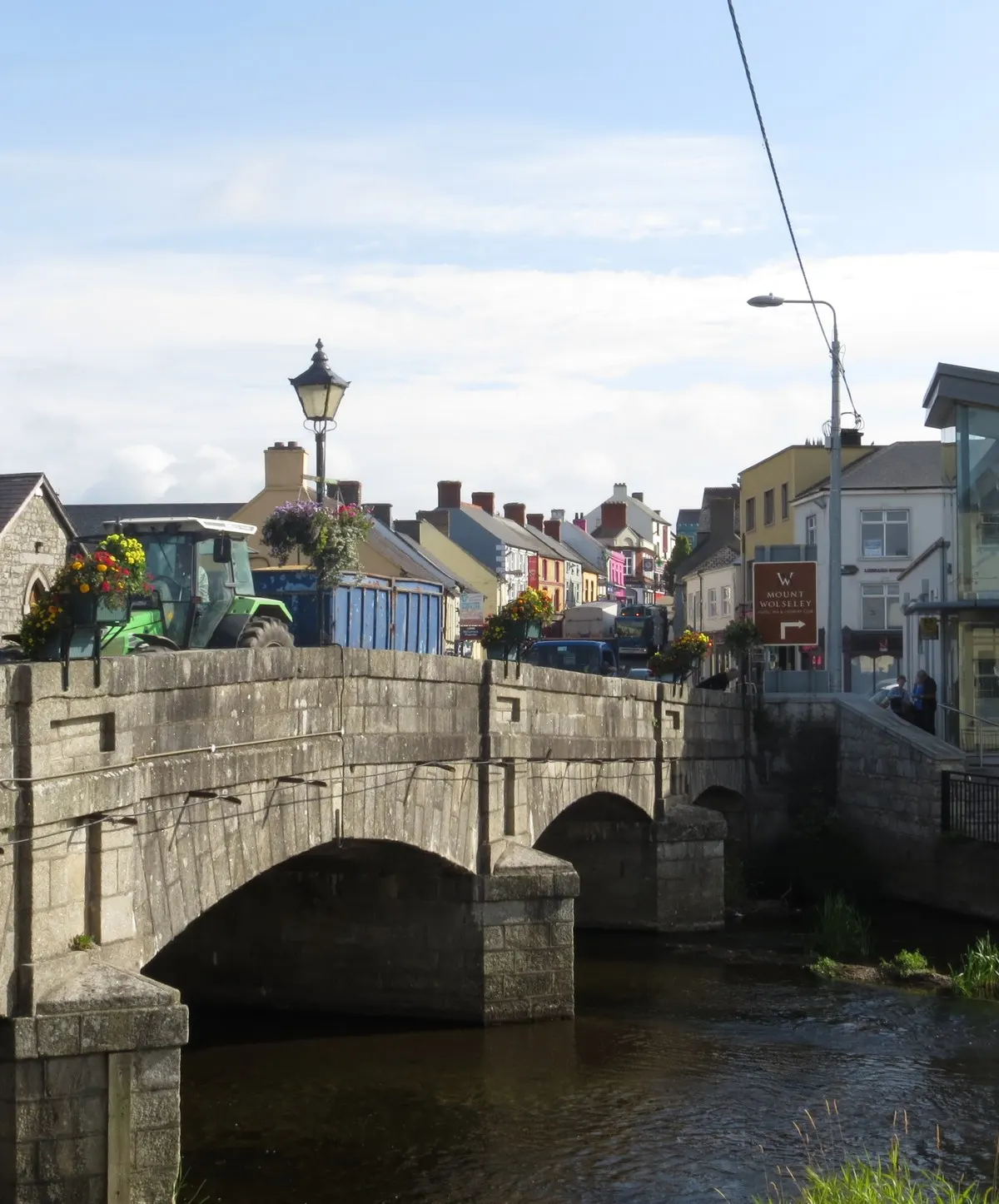 The stone bridge carrying the N81 over the River Slaney in the centre of Tullow, with flower baskets on the parapet and the painted shopfronts of the main street behind