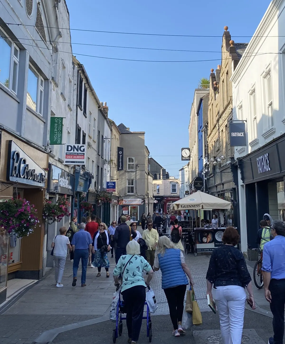 Shoppers walking up the pedestrianised North Main Street in Wexford town on a sunny afternoon, shopfronts and hanging baskets down both sides